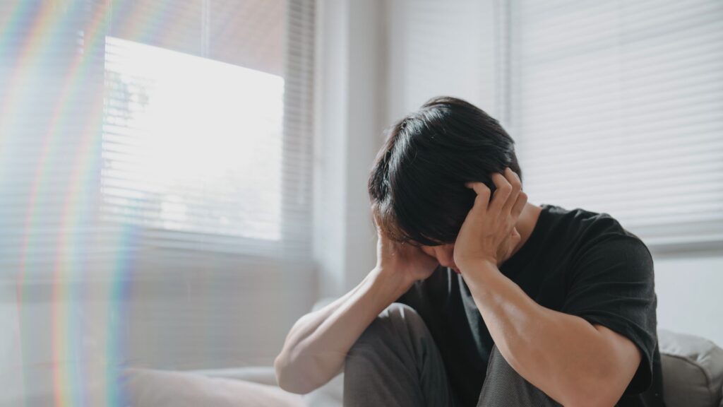 man with head in hands, knees pulled up to chest in an empty, windowed room