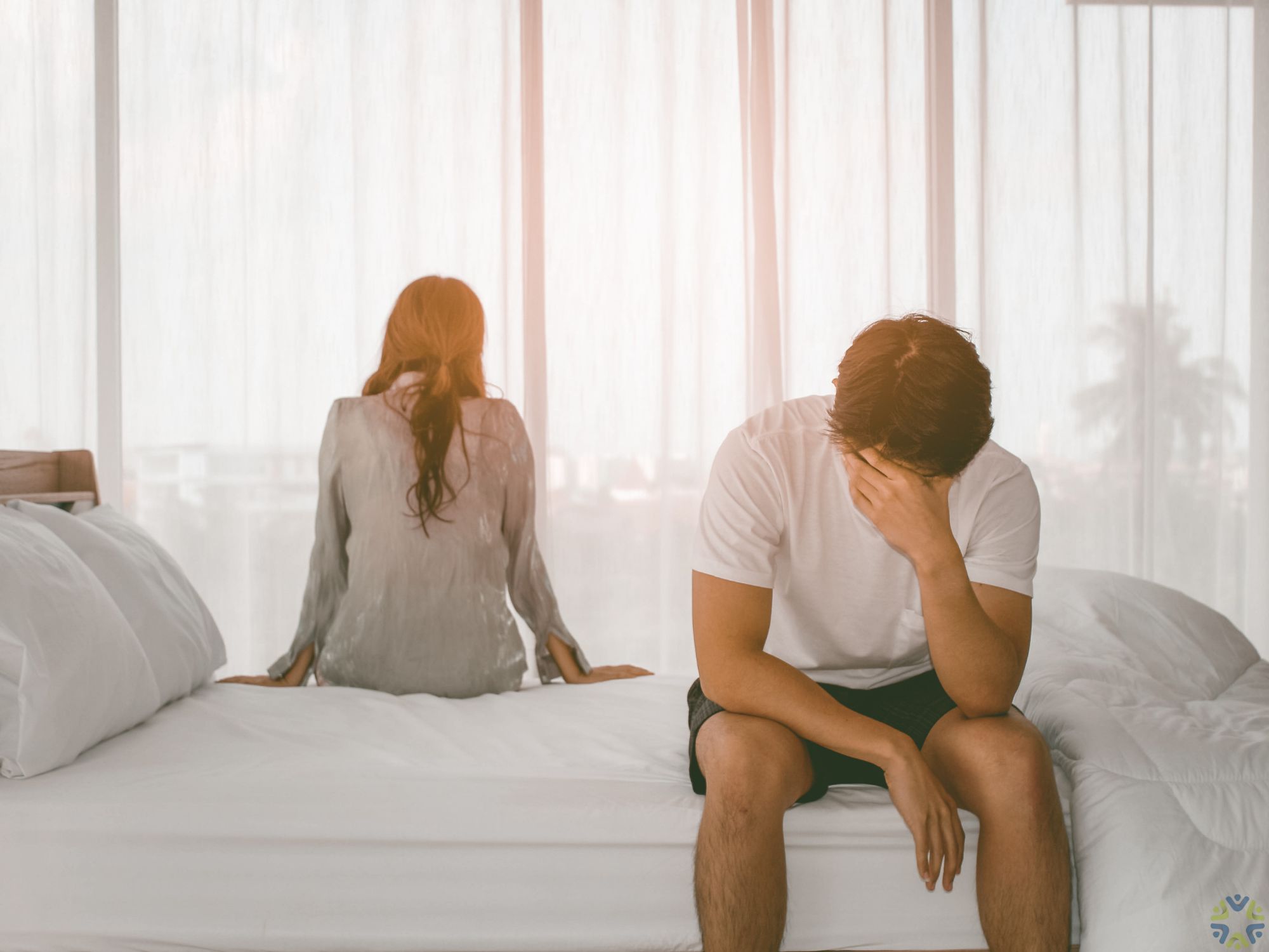 people on opposite sides of a bed, backs to one another, man in foreground with head in hand